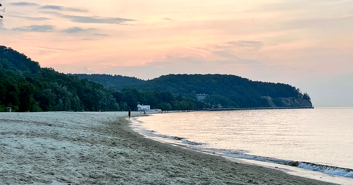 Szeroka piaszczysta plaża nad Zatoką Gdańską o zachodzie słońca, w tle zalesiona skarpa i molo wchodzące w morze
