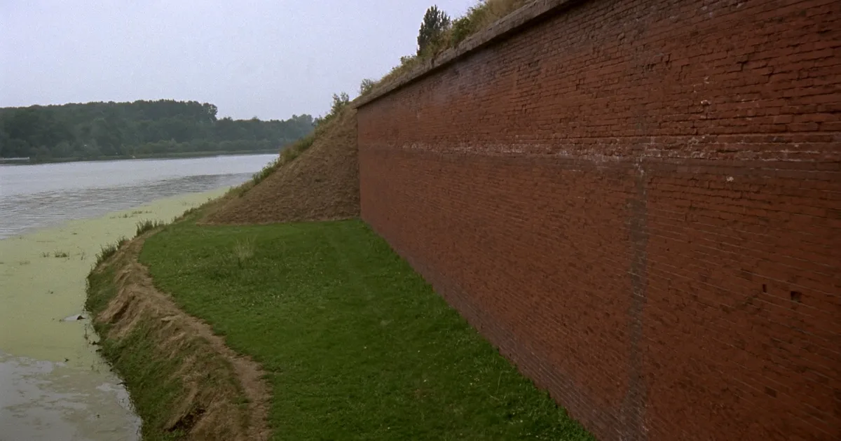 Ceglany mur obronny i trawiasty wał nad spokojną wodą kanału portowego