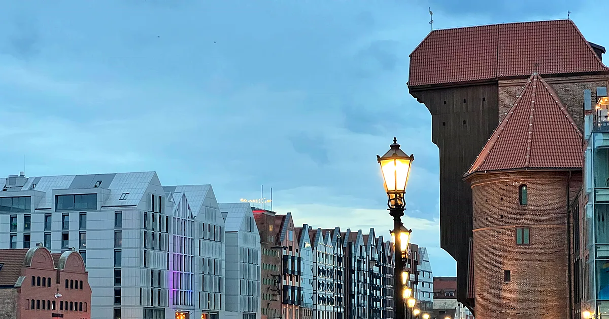 Żuraw nad Motławą o zmierzchu — ceglane wieże i drewniana nadbudowa dźwigu, zapalone latarnie Długiego Pobrzeża i rząd kamienic odbijający się w wodzie